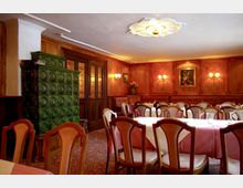 Ein gemütlicher Gastraum mit Holzwänden, gedeckten Tischen und gepolsterten Holzstühlen. Im Raum befinden sich eine grüne Kachelheizung, Wandlampen sowie ein Gemälde und Blumendekorationen.