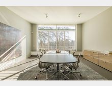 Heller Besprechungsraum mit großem ovalem Tisch und sechs Bürostühlen auf einem Teppich. Große Fenster mit Jalousien und Blick auf Bäume dominieren die Rückwand, während links ein abstraktes Kunstwerk hängt und rechts ein Sideboard steht.