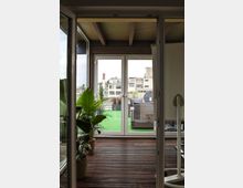 Blick durch eine Glastür auf eine moderne Terrasse mit Holzboden, grüner Kunstrasenfläche und Rattanmöbeln; links stehen große Topfpflanzen. Im Hintergrund sind Wohngebäude zu sehen.