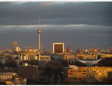 Panorama von Berlin mit Blick auf den Fernsehturm, das moderne Regierungsviertel und den Reichstag mit seiner gläsernen Kuppel. Im Vordergrund sind verschiedene Gebäude und Dächer sichtbar, während der Himmel von dunklen Wolken bedeckt ist.