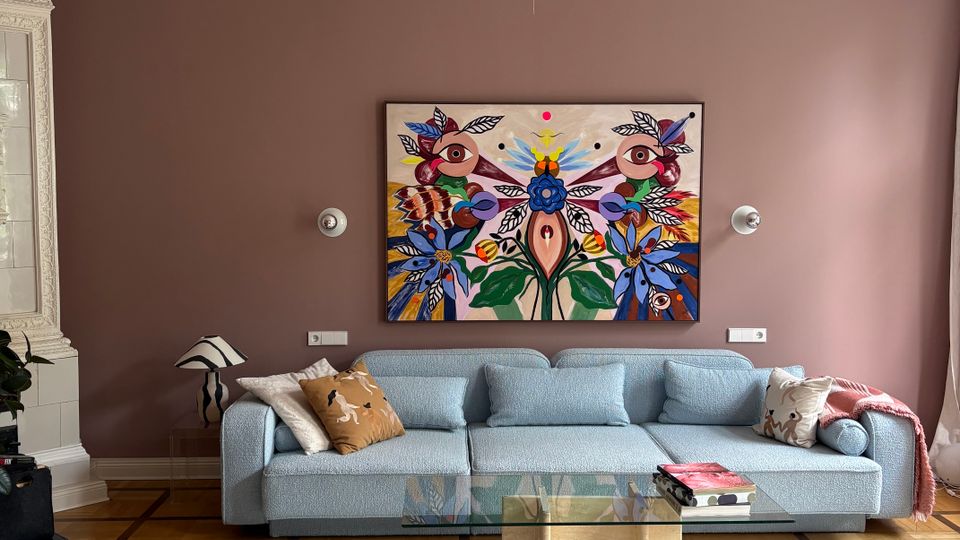 Elegantes Wohnzimmer mit hohen Decken, reich verzierten Stuckleisten und einer großen runden Pendelleuchte. Vor einer mauvefarbenen Wand steht ein hellblaues Sofa unter einem farbenfrohen Wandbild; davor liegen ein Glas-Couchtisch auf hellem Teppich und Parkettboden.