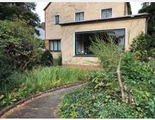 Zweistöckiges Wohnhaus mit beiger Fassade und großen Fenstern, umgeben von einem verwilderten Garten mit dichtem Bewuchs aus Gras, Sträuchern und Efeu. Ein gepflasterter Weg führt durch den Vordergarten.