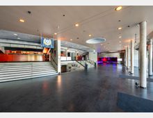 Großer, moderner Innenraum mit Betonwänden, mehreren Treppen und Säulen. Im Hintergrund befinden sich Bereiche mit roten Akzenten und beleuchtete Bühnenvorhänge, während die Decke runde Lichtöffnungen aufweist.