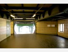 Ein Durchgangsbereich unter einer Brücke mit gelb-beigen Fliesenwänden und einer dunkelgrauen Bodenfläche. Die Struktur der Brücke besteht aus grünen Stahlträgern, und Tageslicht sowie Fahrräder sind am Ausgang sichtbar.