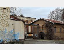 Verlassener Industriehof mit teilweise verputzten Backsteingebäuden, abblätternder Fassade und Graffiti an einer Seitenwand. Im Hintergrund stehen flache Hallen mit rostigem Blechdach, schmalen Fenstern, überwucherten Sträuchern und kahlen Bäumen.