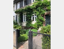 Zweistöckiges Wohnhaus mit weißen Fenstern und schwarzen Fensterläden, teilweise von Kletterpflanzen umrankt. Der Vorderbereich umfasst einen kleinen Garten mit gepflegten Büschen, Pflastersteinen und einem schwarzen Metallzaun mit Tor.