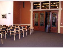 Ein Konferenzraum mit mehreren Reihen von Holzstühlen mit gepolsterten Sitz- und Rückenlehnen. Im Hintergrund befinden sich geöffnete Holztüren mit Ornamentverglasung, die in einen angrenzenden Raum führen, sowie Wände mit roten Backsteinelementen und dezentem Dekor an der Seite.