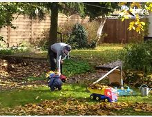 Ein herbstlicher Garten mit einer Wiese, die mit Laub bedeckt ist. Eine erwachsene Person und ein Kind spielen oder arbeiten, während Kinder-Spielzeug, ein roter Trettraktor und eine gelbe Spielzeug-Lkw-Schaufel sichtbar sind; im Hintergrund stehen Bäume, ein Holzzaun und ein Geräteschuppen.