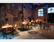 Industrieller Innenraum mit Sichtmauerwerk und hohen Decken, eingerichtet mit einfachen Holztischen und Stühlen. An den Tischen stehen schlichte Dekorationen, während Pendelleuchten von der Decke hängen, durch die Fenster fällt gedämpftes Tageslicht.