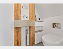 Modernes Badezimmer mit minimalistischem Design, das schrägen Wänden folgt. Ein Waschbecken aus Beton ist in eine Wand mit sichtbaren Holzträgern integriert, daneben befinden sich ein wandmontiertes WC und weiße Regale.