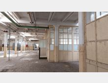 Ehemaliger Industrieinnenraum mit großen Fenstern und teils verglasten Holzwänden, abgenutztem Holzboden und Rohren an der Decke. Der Raum ist leer und von sichtbaren Abnutzungsspuren geprägt.