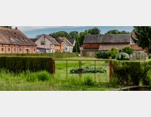 Dorfansicht mit mehreren Häusern aus Backstein und Putz, umgeben von kleinen Gärten mit Hecken, Gewächshäusern und einer Wiese im Vordergrund, die durch einen Metallzaun abgegrenzt ist. Im Hintergrund sind Bäume und eine ländliche Umgebung sichtbar.