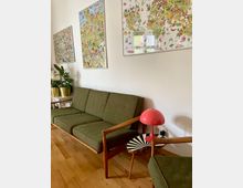 Wohnzimmer mit einer grünen Retro-Couch und einem passenden Sessel auf Holzboden. An der Wand hängen bunte Poster mit Weltkarten-Motiven, daneben ein Beistelltisch mit einer auffälligen rot-weißen Lampe.
