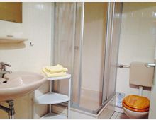 Ein kleines Badezimmer mit beiger Keramik und Fliesen. Es enthält eine Duschkabine, ein Waschbecken mit Spiegel, eine weiße Eckablage mit gelben Handtüchern und eine Toilette mit einem WC-Sitz aus Holz.