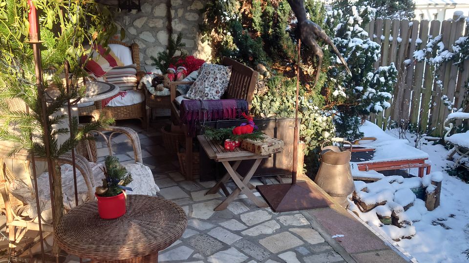 Überdachte Terrasse mit gefliestem Steinboden, Sitzmöbeln aus Rattan und Holztischen, dekoriert mit Pflanzen und roten Ornamenten. Im Hintergrund ein verschneiter Garten mit Holzzaun und immergrünen Bäumen.
