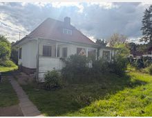 Einfamilienhaus mit weißer Putzfassade und rotem Ziegeldach, mit Wintergartenanbau und kleinen Dachfenstern. Davor liegt ein verwilderter Garten mit hohem Gras und Sträuchern, ein schmaler Gehweg führt am Haus entlang; im Hintergrund stehen Bäume unter bewölktem Himmel.