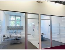 Moderne Büroumgebung mit zwei verglasten Besprechungsräumen, getrennt durch transparente Glaswände mit gestreiftem Dekormuster. Im Hintergrund ein großes Fenster mit Blick ins Grüne und im Innenraum ein Tisch sowie ein Whiteboard an der Wand.