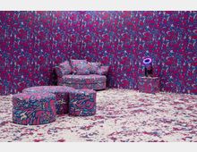 Wohnzimmer mit auffälligem, einheitlichem Design in lebhaften Farben. Wände, Sofa, Hocker und Teppich sind mit einem abstrakten, pink-blauen Muster bedeckt; daneben steht ein kleiner, mit demselben Muster verzierter Beistelltisch mit einer Lampe.