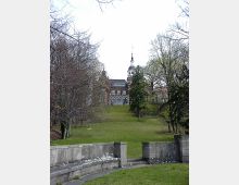 Historisches Gebäude mit Zierturm im Barockstil, umgeben von einer parkartigen Grünfläche mit Rasen, Bäumen und einer Steinbalustrade im Vordergrund. Der Himmel ist bewölkt, und der Blick führt leicht bergauf zum Gebäude.