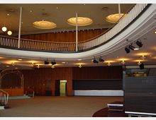 Großer Innenraum mit geschwungener Galerie, Holzwänden und runden Deckenleuchten. Der Raum verfügt über eine Bühne, Deckenstrahler und eine Treppe, die zur Galerie führt.