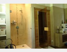 Modernes Badezimmer mit begehbarer Dusche und angrenzender Sauna mit Holztür. Links sind offene weiße Regale mit geflochtenen Körben, rechts dekorative Elemente und ein Sideboard mit Kerzenhalter.