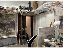 Vollgestellter Atelier- bzw. Lagerraum mit großen gerollten Leinwänden und Verpackungsmaterial auf hohen Regalen, einem großen Bild an der linken Wand und weißen Einbauschränken rechts. Im Vordergrund stehen Arbeitsutensilien wie Farben, Pinsel und Behälter auf einem Tisch; hinten führt eine offene Tür in einen schmalen Nebenraum.