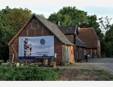 Historisches Gebäudeensemble aus rotem Backstein mit Fachwerkelementen, umgeben von Bäumen und mit einem gepflasterten Hof. An der Fassade eines Gebäudes hängt ein großes Banner der „Deutschen Stiftung Denkmalschutz“ mit der Aufschrift „Wir bauen auf Kultur. Ihre Spende hilft!“.