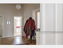 Heller Flur in einer Wohnung mit Holzboden und weißen Wänden, ausgestattet mit einer Garderobe voller Jacken auf einem Ständer. Im Hintergrund ist ein Raum mit einem roten Barhocker und großen Fenstern erkennbar.