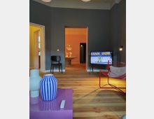 Wohnzimmer mit Holzboden, minimaler Einrichtung und moderner Dekoration. Im Vordergrund steht ein lila Tisch mit Vasen, rechts ein roter Stuhl, und im Hintergrund ist ein Flur mit warmem Licht und einer hängenden Leuchte zu sehen, sowie ein eingeschalteter Fernseher auf der rechten Seite.