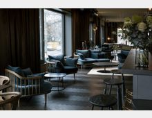 Lounge-Bereich mit modernen, dunkelblauen Polstermöbeln und Holzrahmen, runden Beistelltischen und großen Fensterfronten, durch die natürliche Beleuchtung einfällt. Die Einrichtung umfasst dunklen Holzboden, Vorhänge und stilvolle Dekoration, darunter eine Vase mit Blumen auf der Theke.