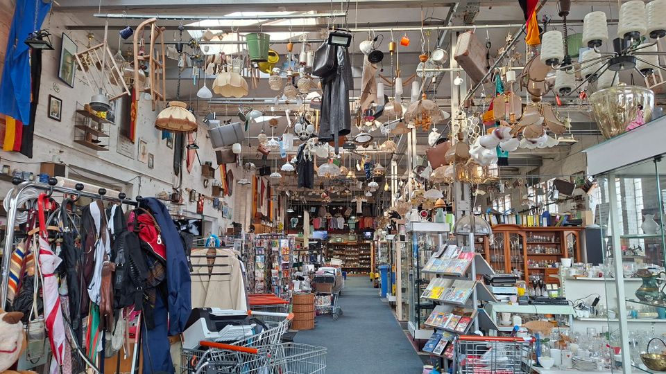 Innenansicht einer großen, hallenartigen Secondhand- und Trödlerladenfläche mit hohen Decken, dicht bestückten Regalen und Vitrinen. Von der Decke hängen zahlreiche Lampen, Haushaltsgegenstände und Dekorationen; entlang eines Mittelgangs stehen Kleiderständer, Einkaufswagen und Warenstände.