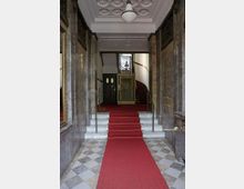 Eingangsbereich eines historischen Gebäudes mit Marmorböden und einem roten Teppich, der über Stufen zu einem verzierten Aufzug und einer Treppe führt. Die Decke ist mit dekorativen Stuckelementen versehen, und links und rechts befinden sich große Spiegel.