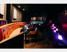 Dunkler Bar- oder Lounge-Innenraum mit einer langen, gepolsterten Lederbank, kleinen runden Tischen und Sesseln. Gedimmte Deckenbeleuchtung und mehrere farbige Tischlampen sorgen für Akzente, dazu Backsteinwand und schwere Vorhänge.