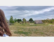 Eine Wiese mit einem einfachen Spielplatz aus Holz, bestehend aus einem Turm mit Rutsche und Klettergerüst, daneben eine kleine, mit Strohdach bedeckte Hütte. Im Hintergrund befindet sich eine grüne Landschaft mit Bäumen unter bewölktem Himmel.