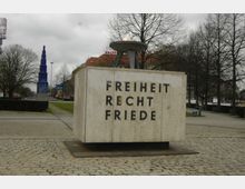 Steinmonument mit der Inschrift „Freiheit, Recht, Friede“ und einer brennenden Flamme darauf, umgeben von einem gepflasterten Platz mit Bäumen; im Hintergrund ist eine moderne, blaue Glasskulptur zu sehen.