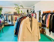 Ein Second-Hand-Laden mit Kleiderständern voller bunter Kleidung, darunter Jacken, Hosen und Oberteile. Im Hintergrund stehen Pflanzen, Dekorationsgegenstände und Regale mit Vintage-Radios.