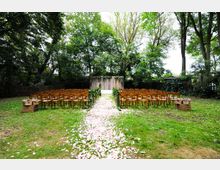 Außenbereich mit einer Hochzeitszeremonie auf einer Wiese, umgeben von Bäumen. Reihen von Holzstühlen flankieren einen mit Blütenblättern gesäumten Gang, der zu einem hölzernen Traubogen mit Blumen führt.