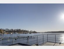 Terrasse mit Geländer überblickt einen ruhigen See mit Stegen und vertäuten Booten. Im Hintergrund sind kahle Bäume und ein klarer Himmel mit strahlender Sonne zu sehen.