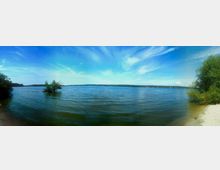 Panoramablick auf einen See mit ruhigem, blauem Wasser, umgeben von Bäumen und Vegetation an den Ufern. Der Himmel ist klar mit leichten Wolken, und im Hintergrund sind kleine Boote sichtbar.