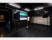 Fotostudio mit schwarzen Wänden und Decke, ausgestattet mit Lichtboxen auf Stativen, einer Schneiderpuppe und mehreren Farbrollen für Hintergrundkulissen. Im Hintergrund ist eine Betonwand sichtbar, links ein abgetrennter Bereich mit Regalbeleuchtung.