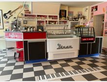 Innenansicht eines Retro-Diners mit schwarz-weißem Schachbrettboden und Verkaufstheke mit Glasvitrinen für Softeis und Kuchen. Hinter der Theke hängen Vintage-Schilder und Regale mit Gläsern und Flaschen; an der Wand ist eine E‑Gitarre befestigt.