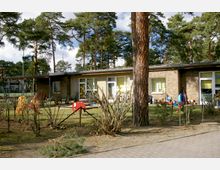 Ein eingeschossiges Gebäude mit Flachdach, umgeben von einem umzäunten Garten mit Kinderspielgeräten wie Rutschen und Schaukelpferden. Im Hintergrund befinden sich hohe Kiefern und einige Wohngebäude, die auf einen ländlichen oder vorstädtischen Kontext hinweisen.