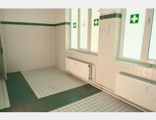 Leerer, gekachelter Raum mit weißer Wandverfliesung und grünem Fliesenstreifen sowie graugrün gemustertem Fliesenboden. An der Fensterwand befinden sich mehrere große Fenster über Heizkörpern; an den Wänden sind grüne Erste-Hilfe-Kreuzsymbole angebracht.