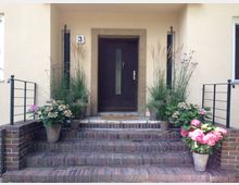 Eingangsbereich eines Hauses mit einer dunklen Holztür und einem schmalen Fenster in einem beigen Putzrahmen. Die Backsteinstufen sind beidseitig mit schwarzen Metallgeländern und zahlreichen Blumentöpfen mit Hortensien und Ziergräsern geschmückt.