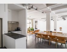 Offener, moderner Innenraum mit einer kleinen Küchenzeile links, einer großen Holztischgruppe mit Stühlen in der Mitte und einer Sitzecke mit Pflanzen und Holzsesseln im Hintergrund. Dahinter liegt ein heller Studiobereich mit weißen Wänden und einem einzelnen Holzstuhl.