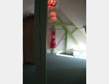 Dachzimmer mit schrägen Wänden, hellgrünen Holzbalken an der Wand und drei bunten Lampen an der Decke. Im Hintergrund ein Holzkleiderschrank und ein hängendes Kinderspielzeug in Form eines rosa Plüschtiers.
