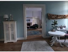 Wohnzimmer mit blauen Wänden und Holzboden, eingerichtet mit weißen Möbeln, darunter ein kleiner runder Tisch und ein Schrank. Im Hintergrund ist ein angrenzendes Zimmer mit Kamin, Teppichboden und Sesseln sichtbar.