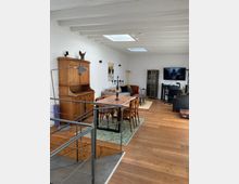 Wohnbereich mit Holzboden und sichtbaren weißen Holzdeckenbalken. Zentral steht ein massiver Esstisch mit Holzstühlen auf einem Teppich, links ein antikes Holzmöbelstück und rechts ein Fernseher auf einem Schreibtisch, daneben eine Vitrine. Im Vordergrund führt ein Metallgeländer zu einer Treppe.