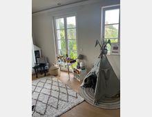 Kinderzimmer mit großen französischen Fenstern, die viel Tageslicht hereinlassen. Der Raum enthält ein graues Tipi-Zelt auf einem runden Teppich, einen Spieltisch, einen Bücherkorb aus Holz und einen gemusterten Teppich auf einem Holzboden.