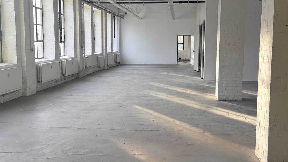 Großer, leerer Industrie-Loft-Raum mit Betonboden, weißen Ziegelstützen und hohen Decken mit sichtbaren Leitungen und langen Deckenleuchten. Links eine Reihe großer Sprossenfenster mit Heizkörpern darunter, durch die Tageslicht in den Raum fällt.